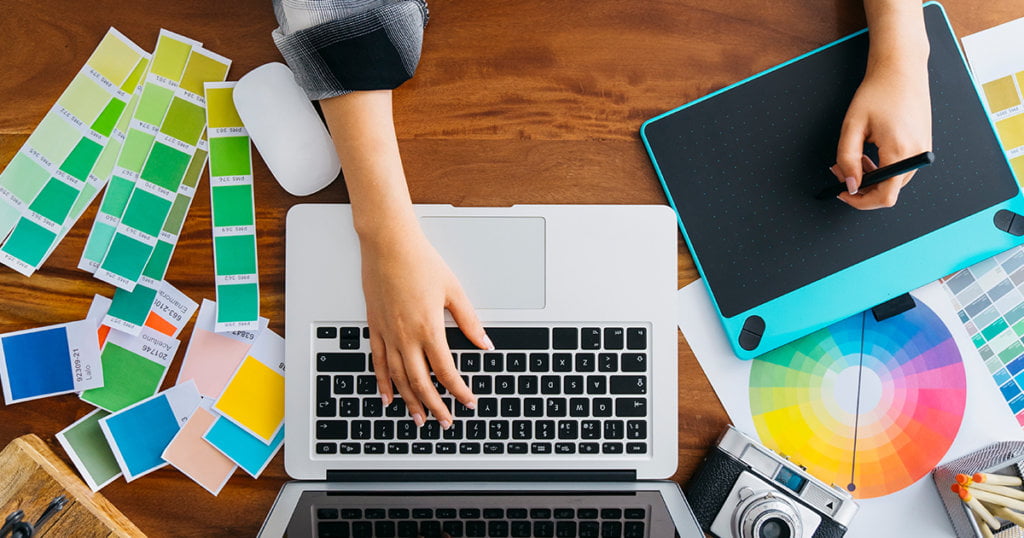 Hand holding a stylus over a drawing pad and another hand hovering the keyboard of a laptop beside a camera and color palettes