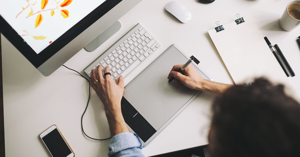 Person holding a stylus pen on top of a drawing pad in front of a desktop computer